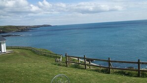 A coastal landscape with a grassy area, a wooden fence, and a white building near the water.