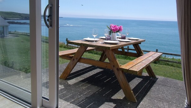 A wooden table set with plates, glasses, and a vase of flowers, overlooking a coastal view.