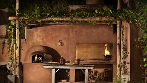 Een buitenoven van baksteen met daarin een vuur, een tafel met servies en een fles, en een rustieke houten pergola met wijnranken.