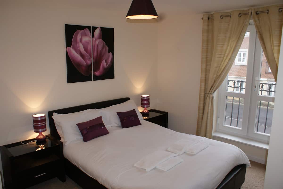 Una cama bien tendida con sábanas blancas y dos almohadas moradas, un cuadro de flores colgado en la pared, dos lámparas de noche y una ventana con cortinas.