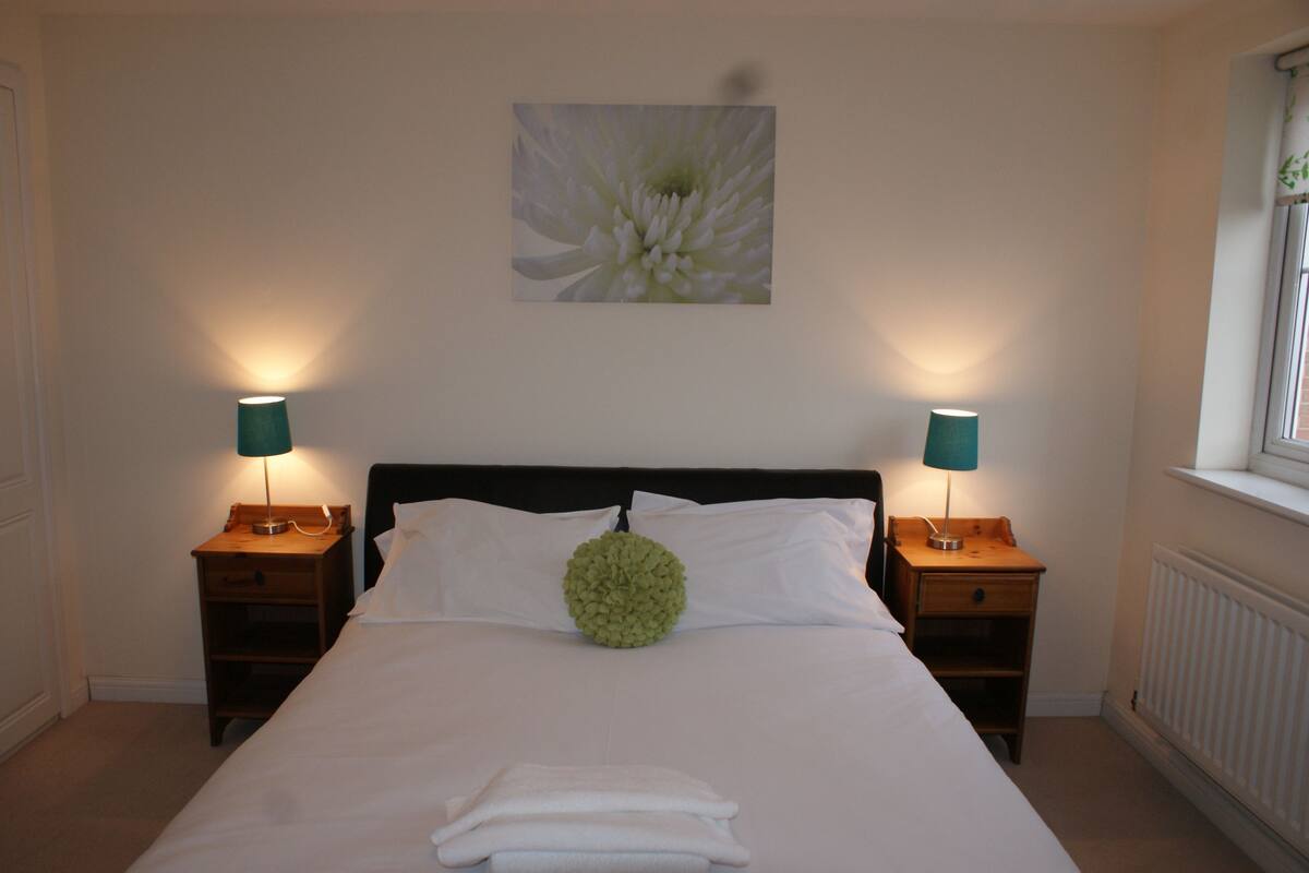 Una cama bien hecha con sábanas blancas, dos mesitas de noche con lámparas, un cojín decorativo verde y un cuadro de flores colgado en la pared.