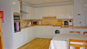 A compact kitchen with white cabinets, a microwave, a washing machine, and a dining table with chairs. - Ornum Cottage (Beauly)
