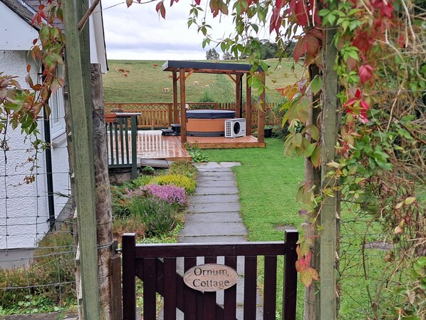 A wooden gate with a sign reading 'Ornum Cottage' leading to a garden area with a wooden deck, a hot tub, and a grassy field in the background. - Ornum Cottage (Beauly)