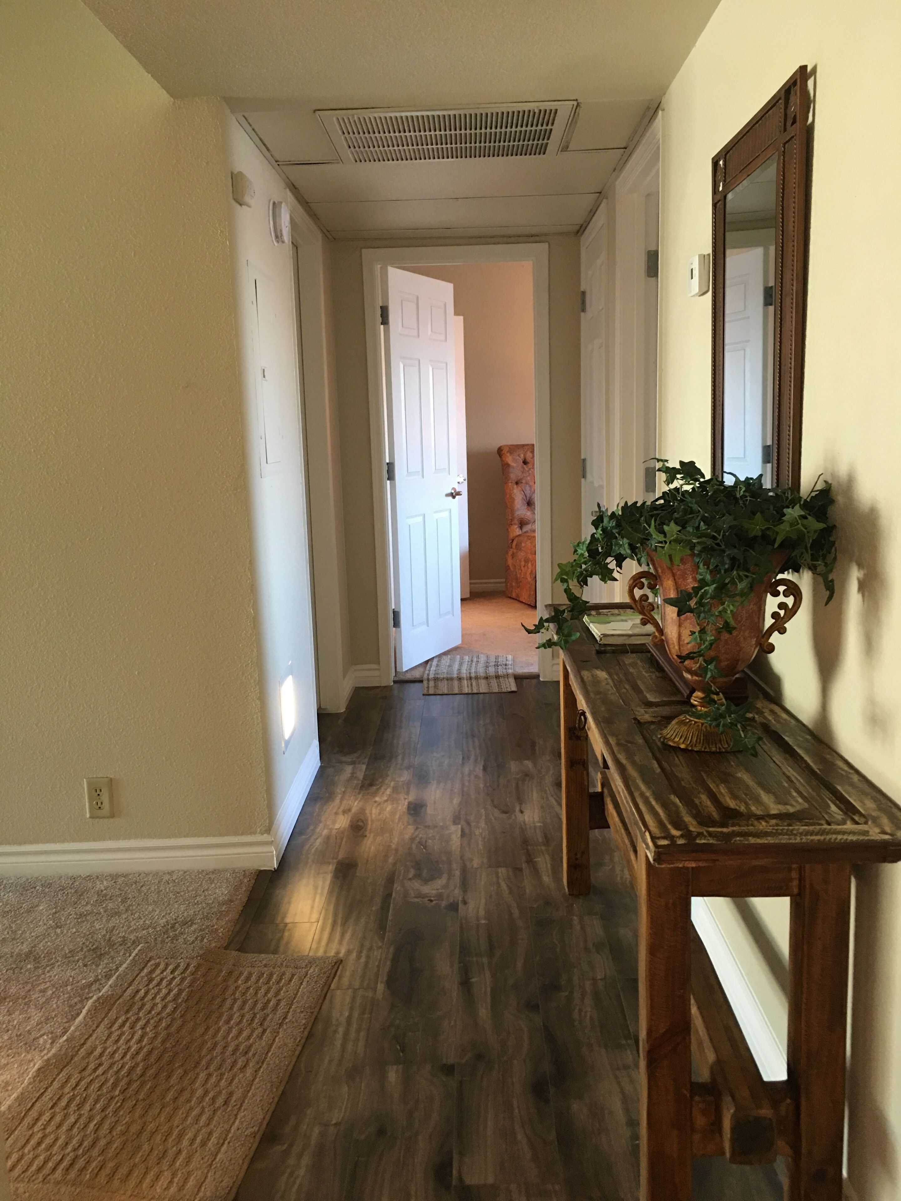 A hallway with wooden flooring, a door at the end, a bench with a plant, and a mirror on the wall.