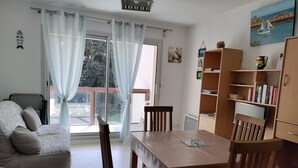 A living room with a sofa, a wooden dining table, chairs, a bookshelf, and a large window with curtains.