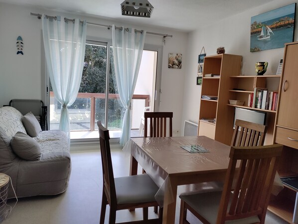 Sala de estar com sofá, mesa de jantar de madeira, cadeiras, estante e uma janela grande com cortinas.