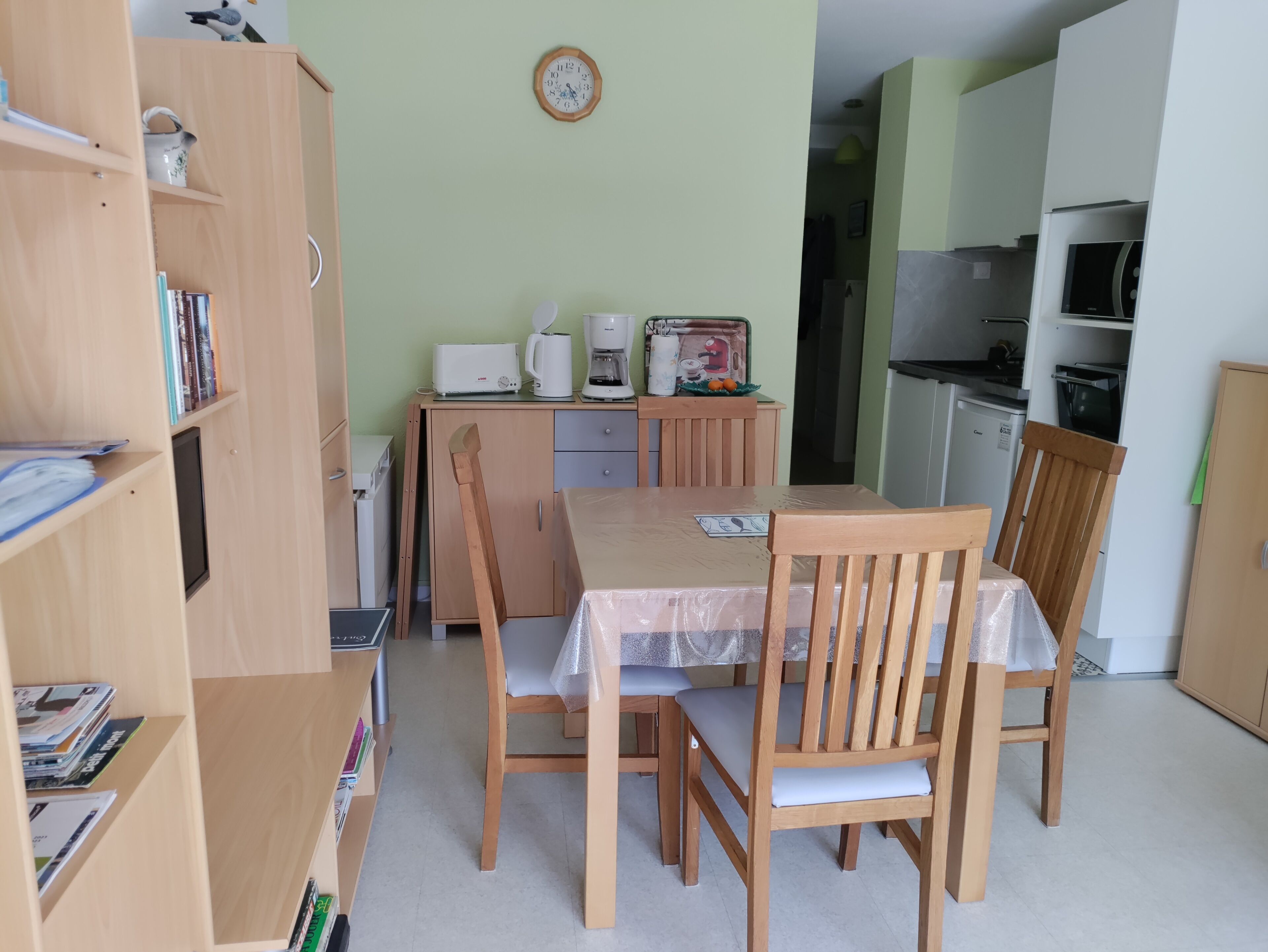 A compact living space with a wooden dining table, chairs, a kitchenette, and a bookshelf.