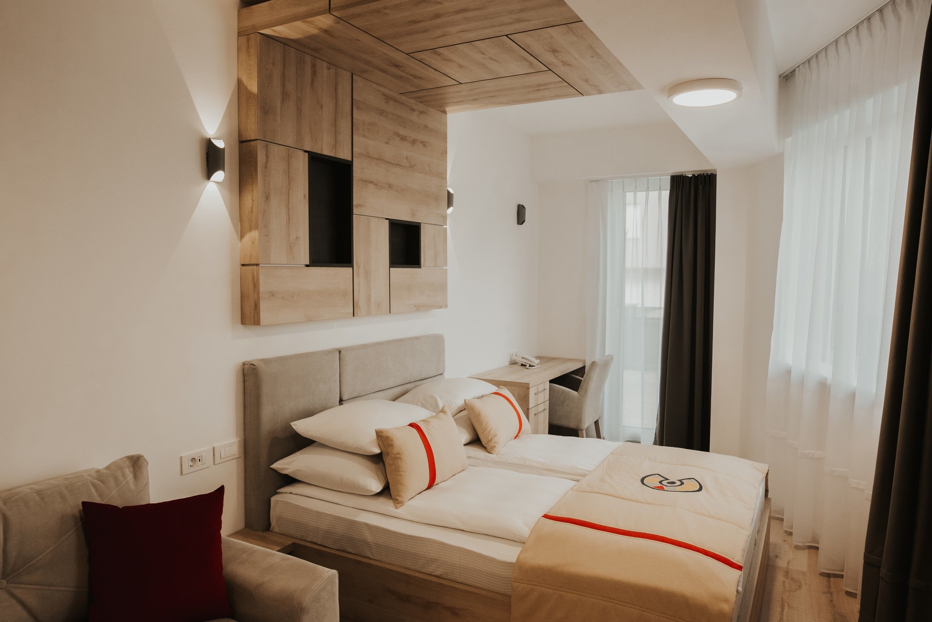 Une chambre d’hôtel moderne avec un plafond en bois, un lit avec une literie beige et des touches de rouge, un bureau et une fenêtre avec des rideaux.