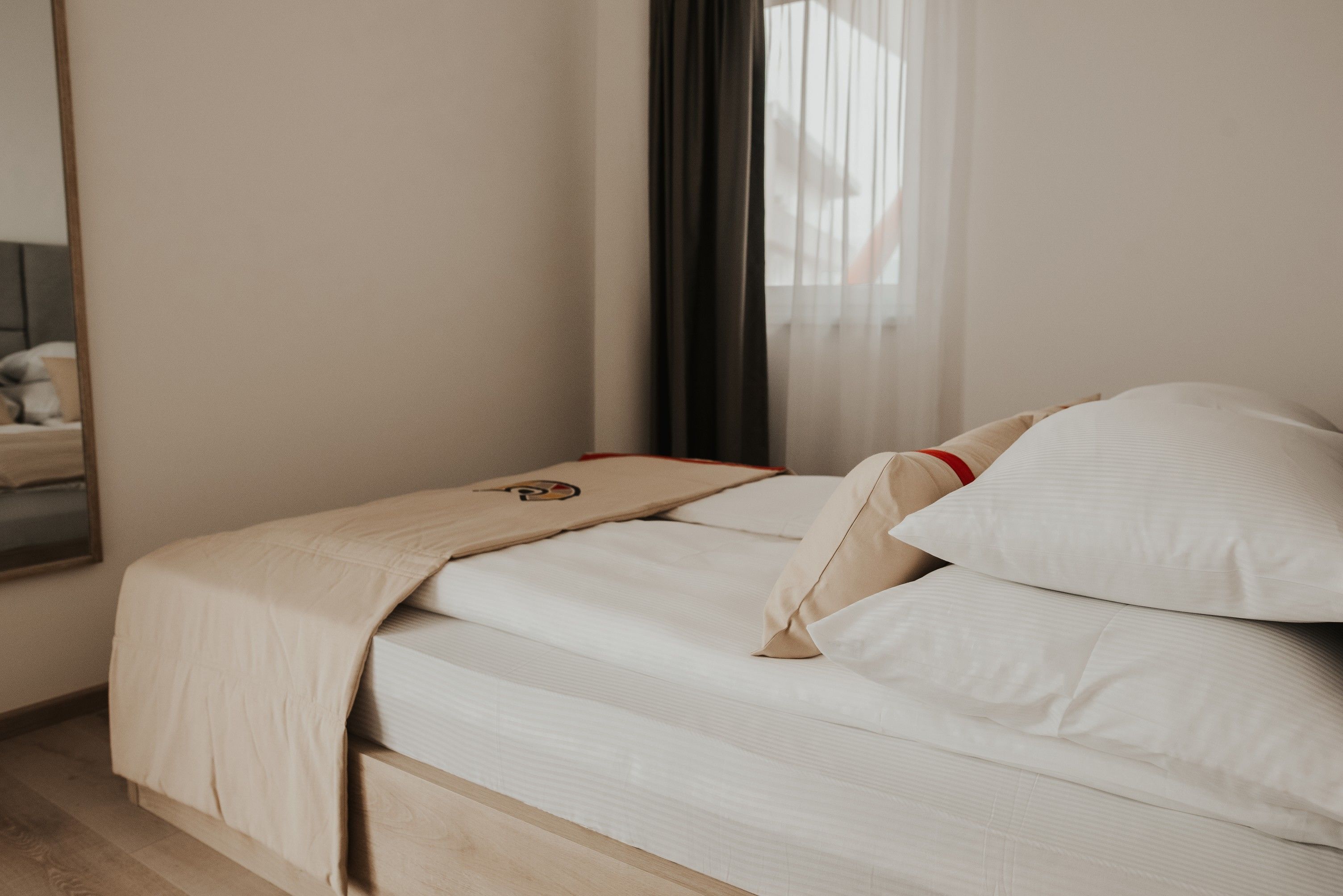 A neatly made bed with a beige blanket and pillows, a wooden headboard, and a mirror on the wall.