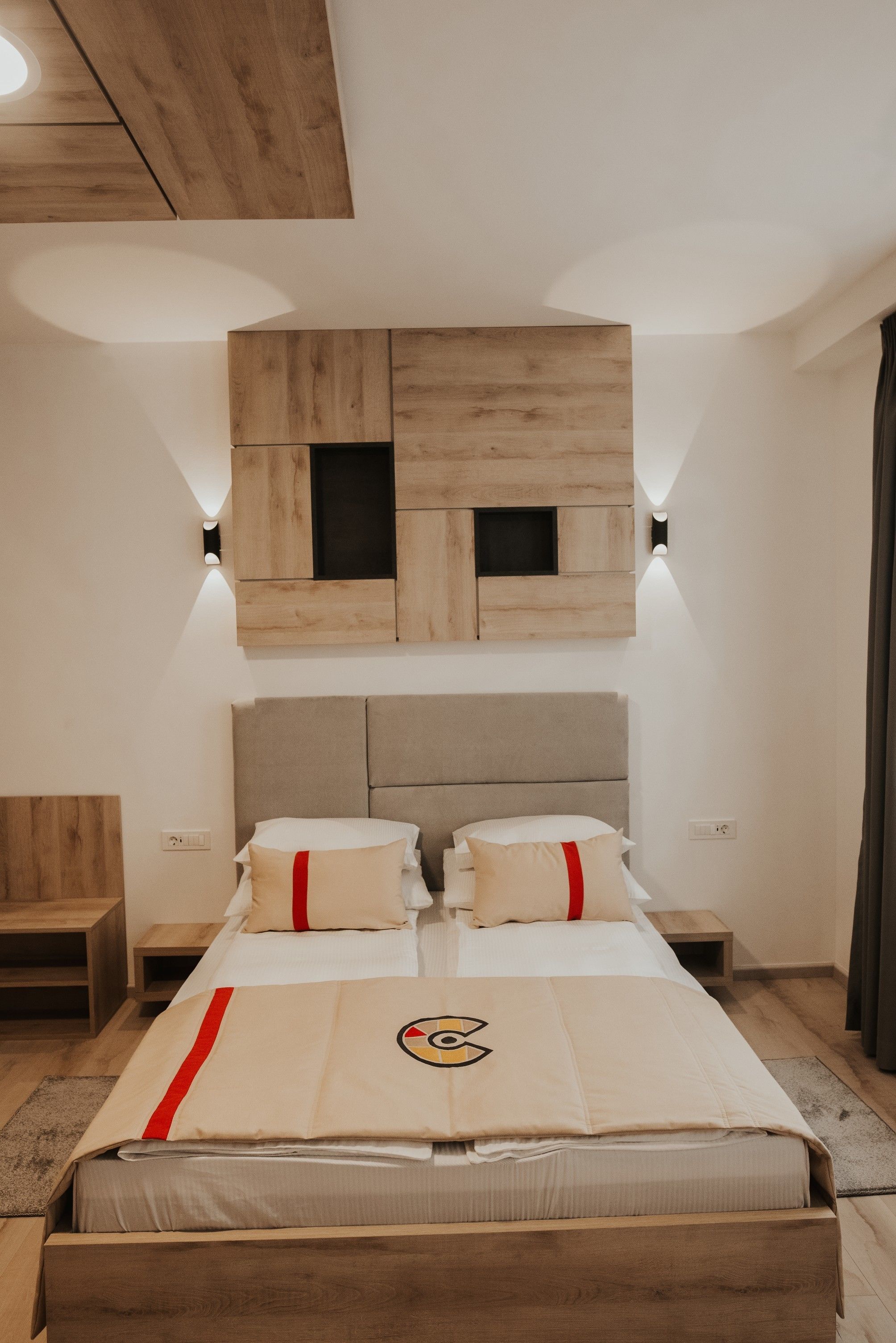 A bedroom with a wooden headboard, two bedside tables, a bed with a beige bedspread and red accents, and built-in wooden shelves above the bed.
