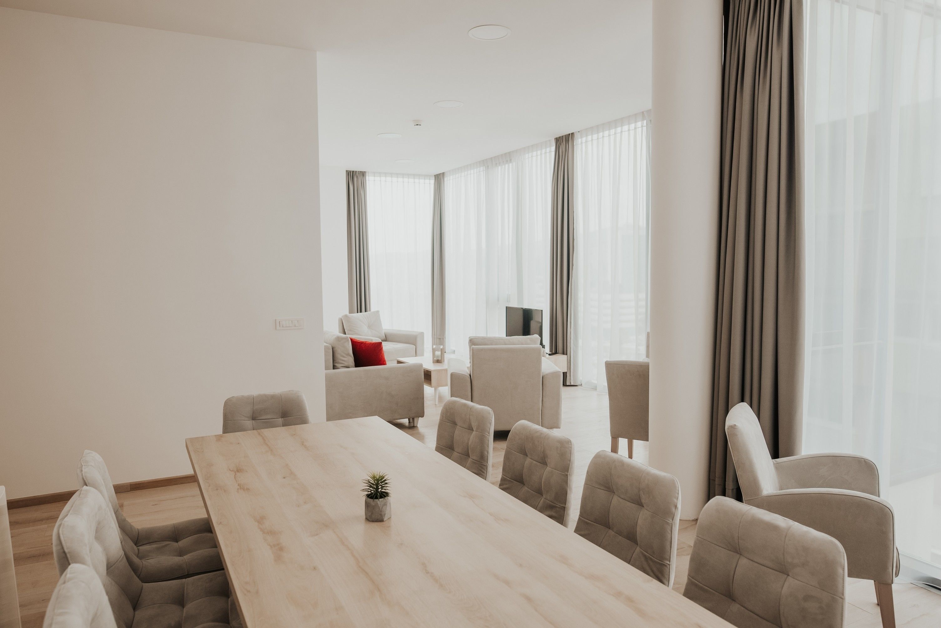Ein modernes Hotelzimmer mit großem Esstisch, bequemen Sitzgelegenheiten und großen Fenstern mit Vorhängen.