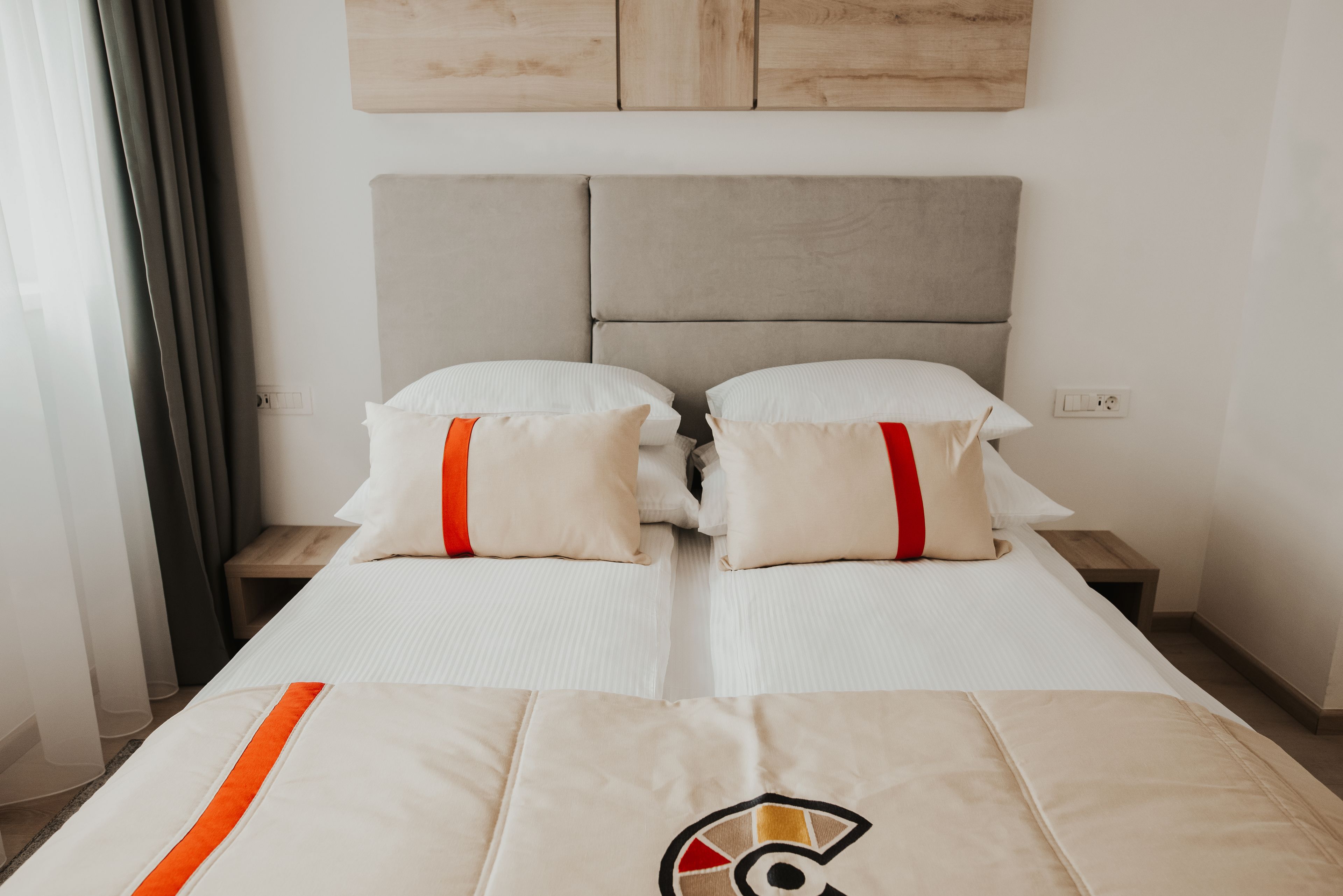 A double bed with white bedding and red accents, a wooden headboard, and a small wooden nightstand beside it.