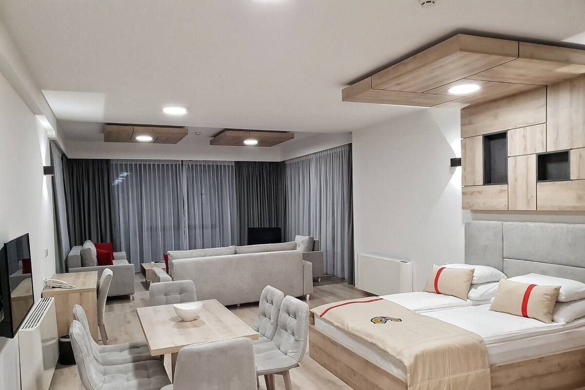 A modern hotel room with a sofa set, a dining table with chairs, a bed with a headboard, a TV, and a window with curtains.
