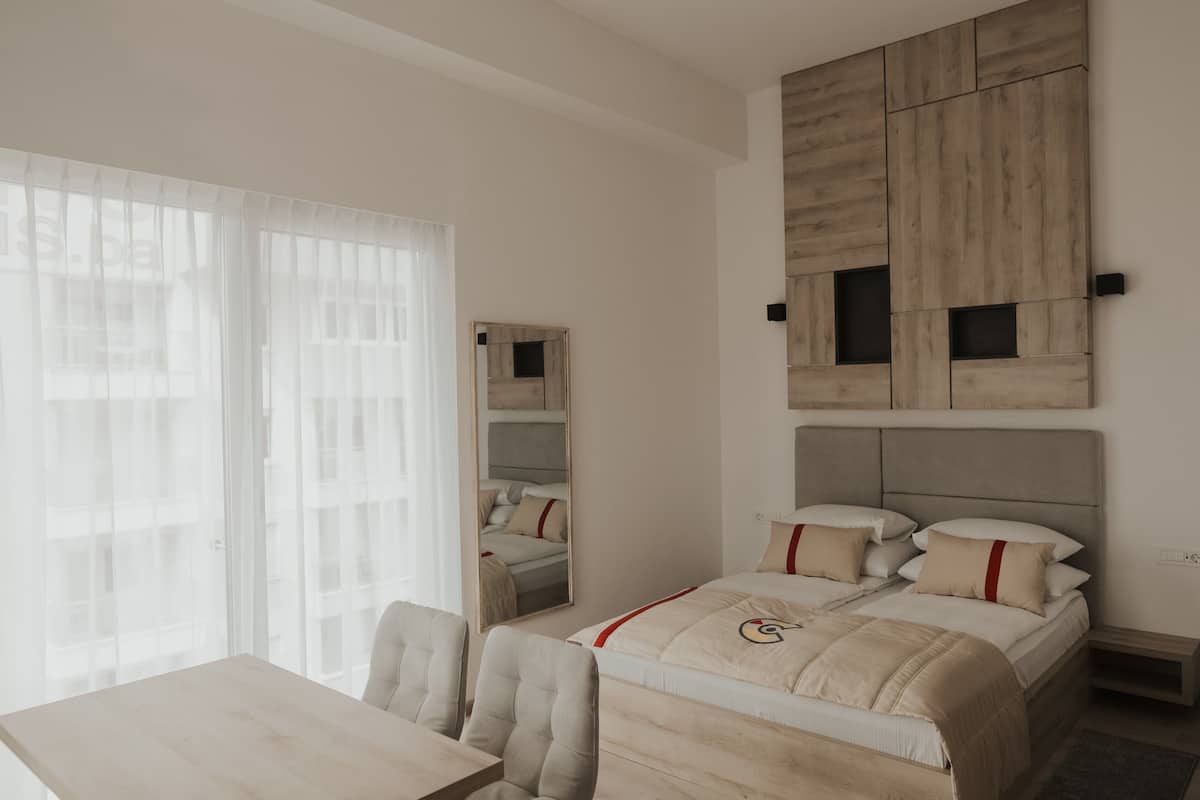 A hotel room with a bed, a desk, and a chair. There is a large mirror on the wall and a window with curtains.