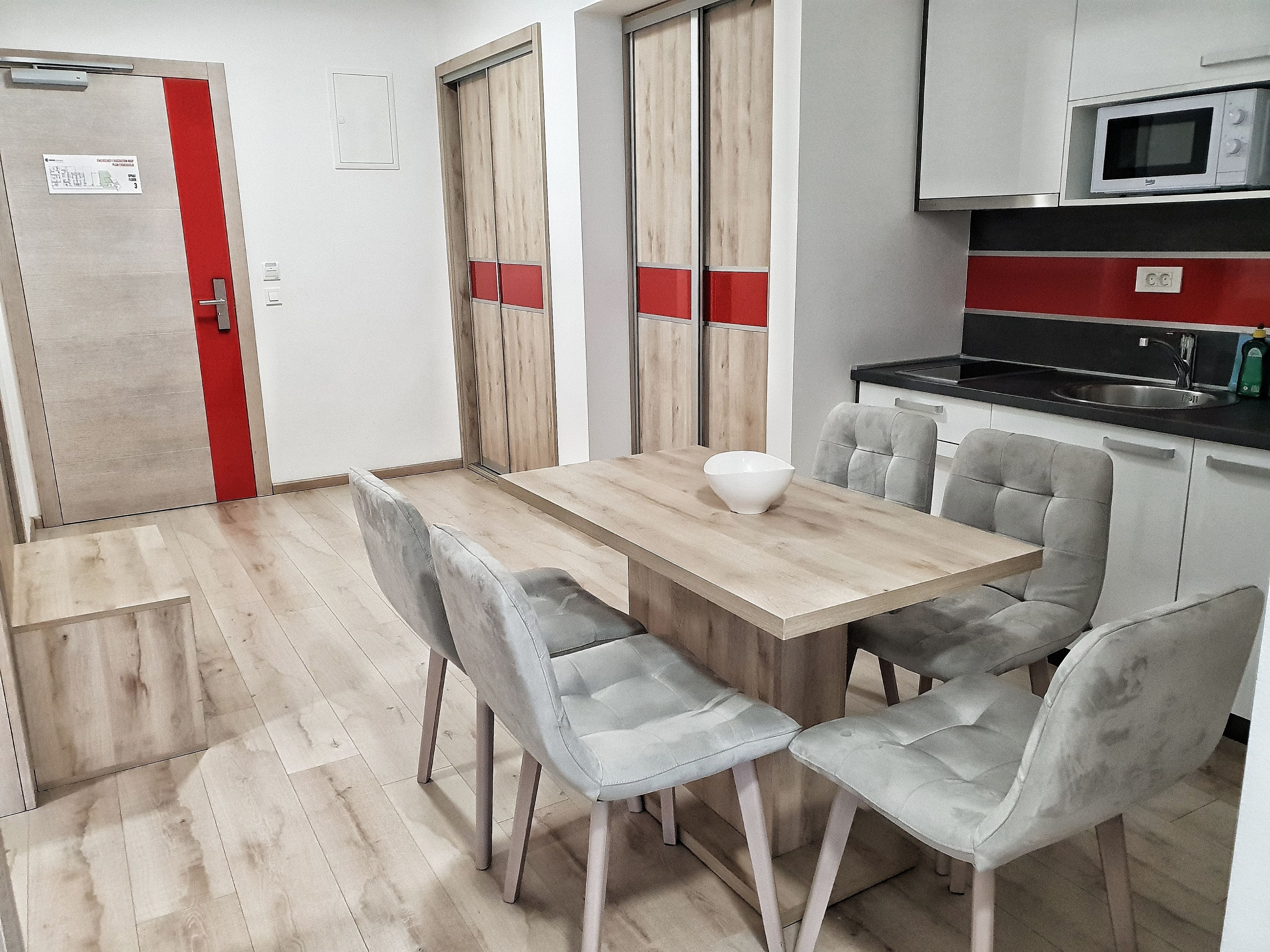 Een moderne keuken met een houten tafel en grijs gestoffeerde stoelen, een magnetron, en een rode deur.