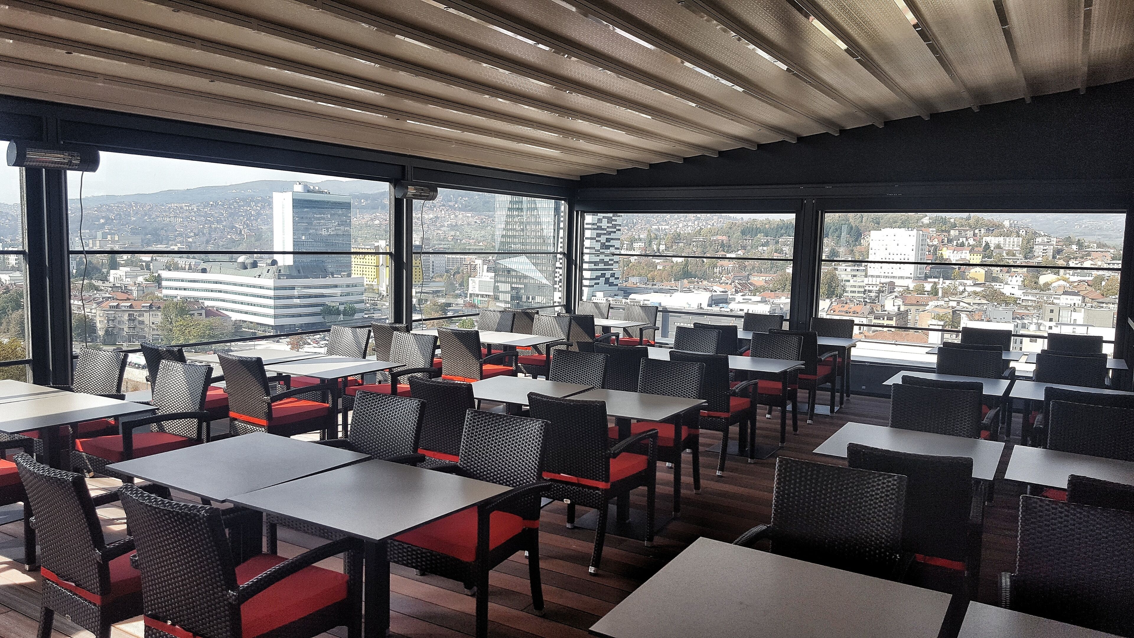A rooftop restaurant with tables and chairs, large windows offering city views.