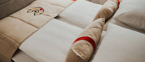 A bed with beige bedding, a pillow with a red band, and a logo on the pillow.