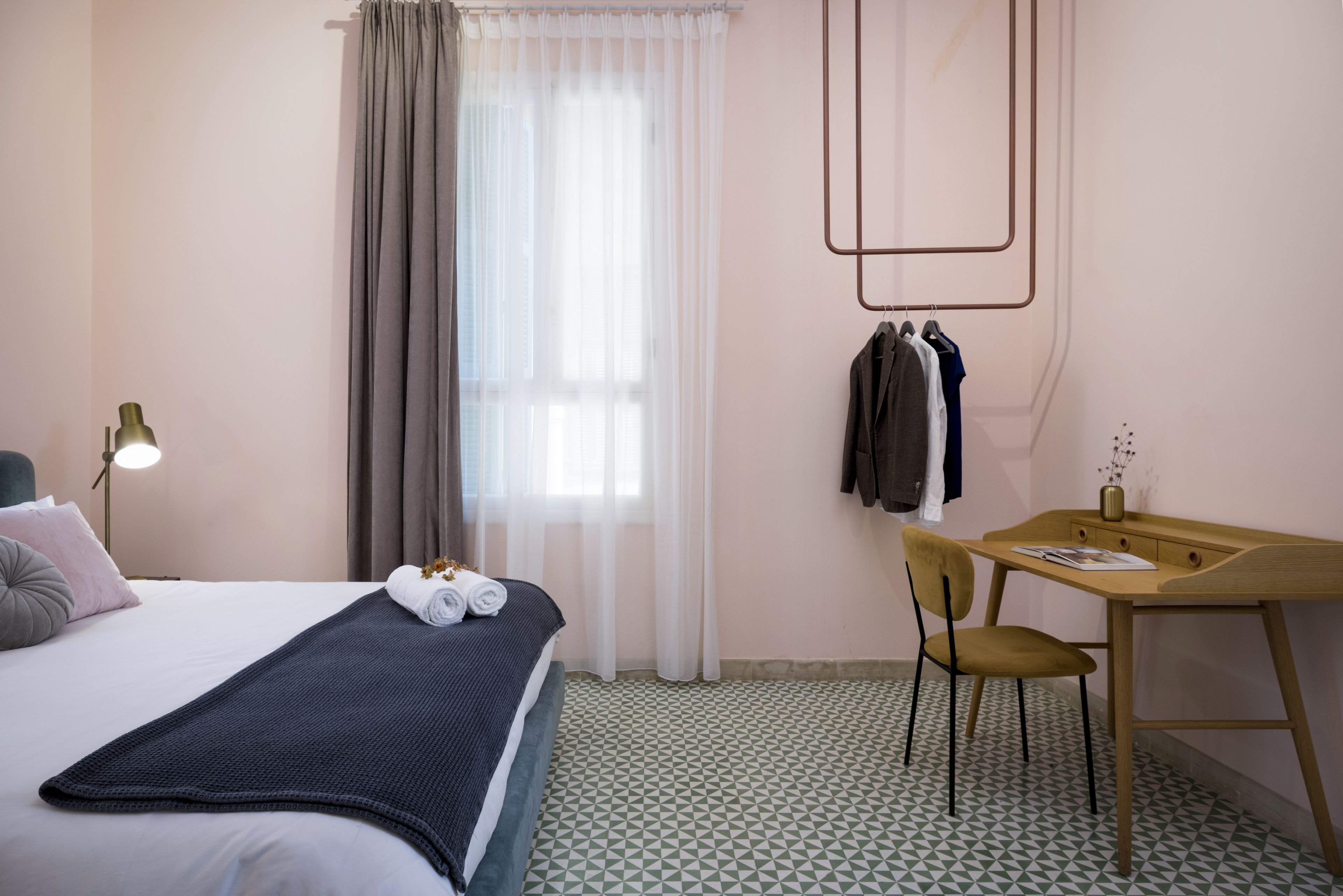 Una habitación de hotel con cama, escritorio, silla, lámpara y ventana con cortinas.