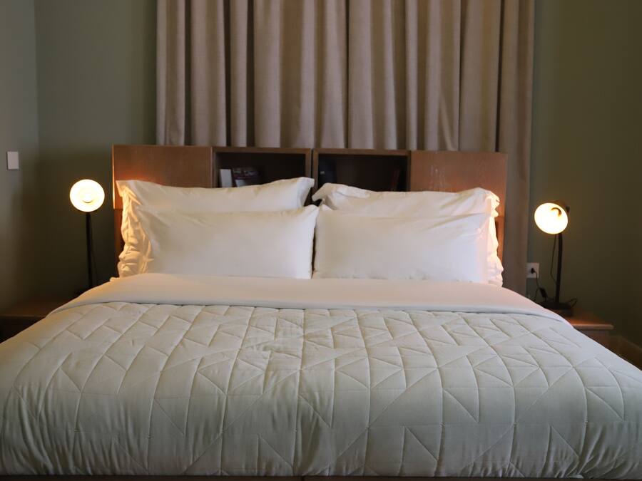 Una cama bien hecha con un edredón blanco acolchado, dos lámparas de noche y un cabecero con estante.