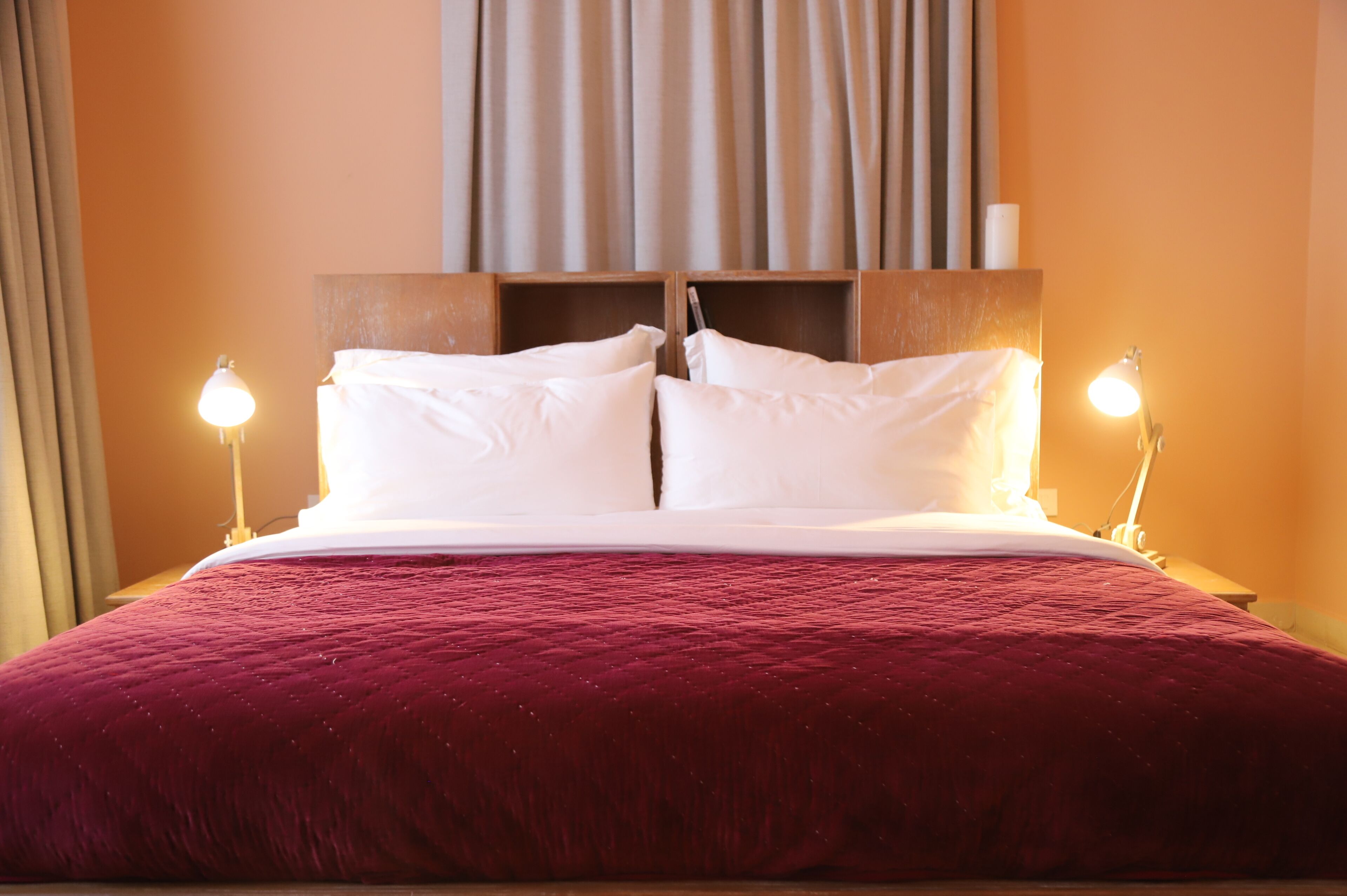 Un lit bien fait, avec du linge de lit blanc et une couverture rouge, de part et d’autre duquel se trouvent deux lampes de chevet.