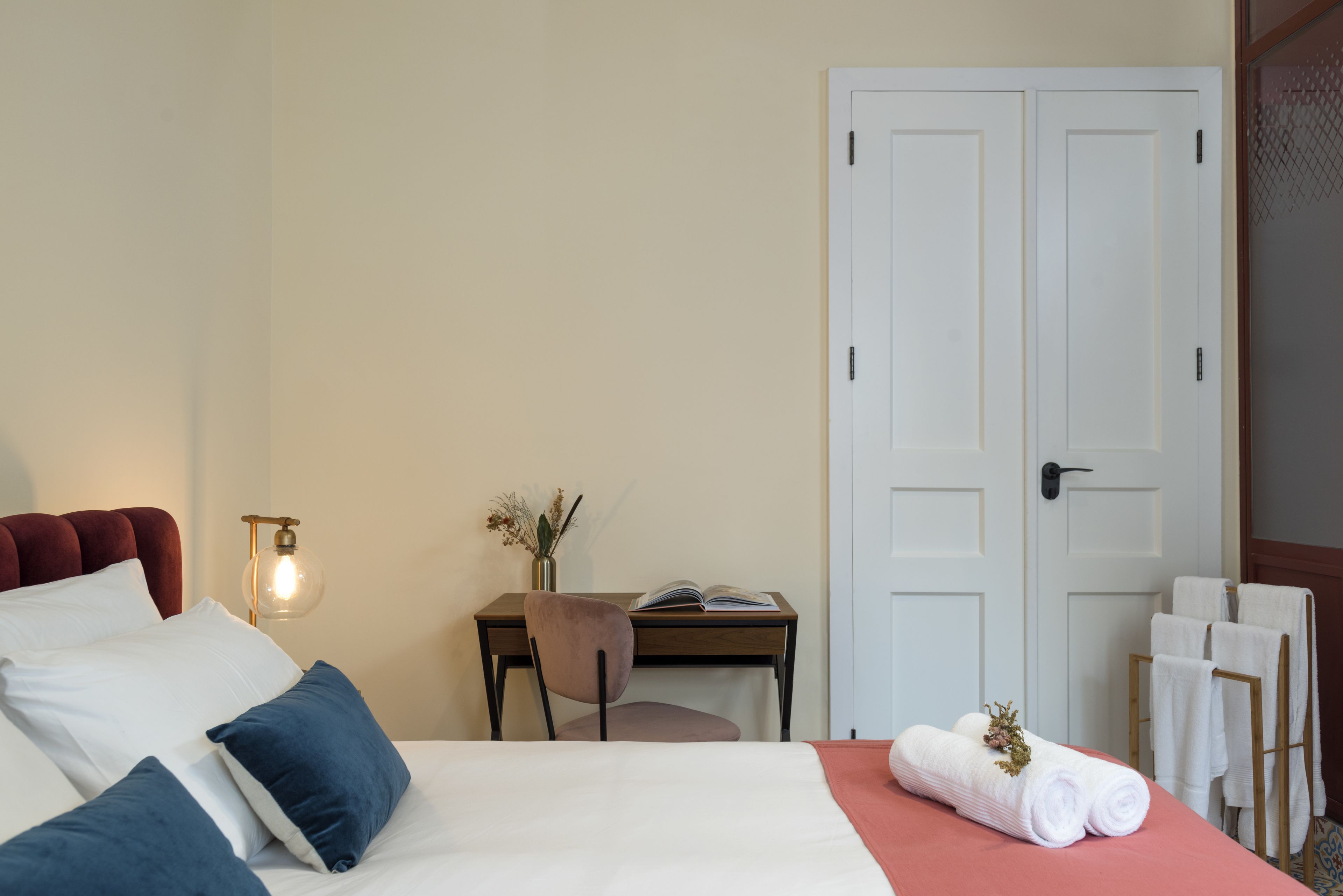 A neatly arranged bedroom with a bed, a desk, and a chair. There is a door and a set of towels on a rack.