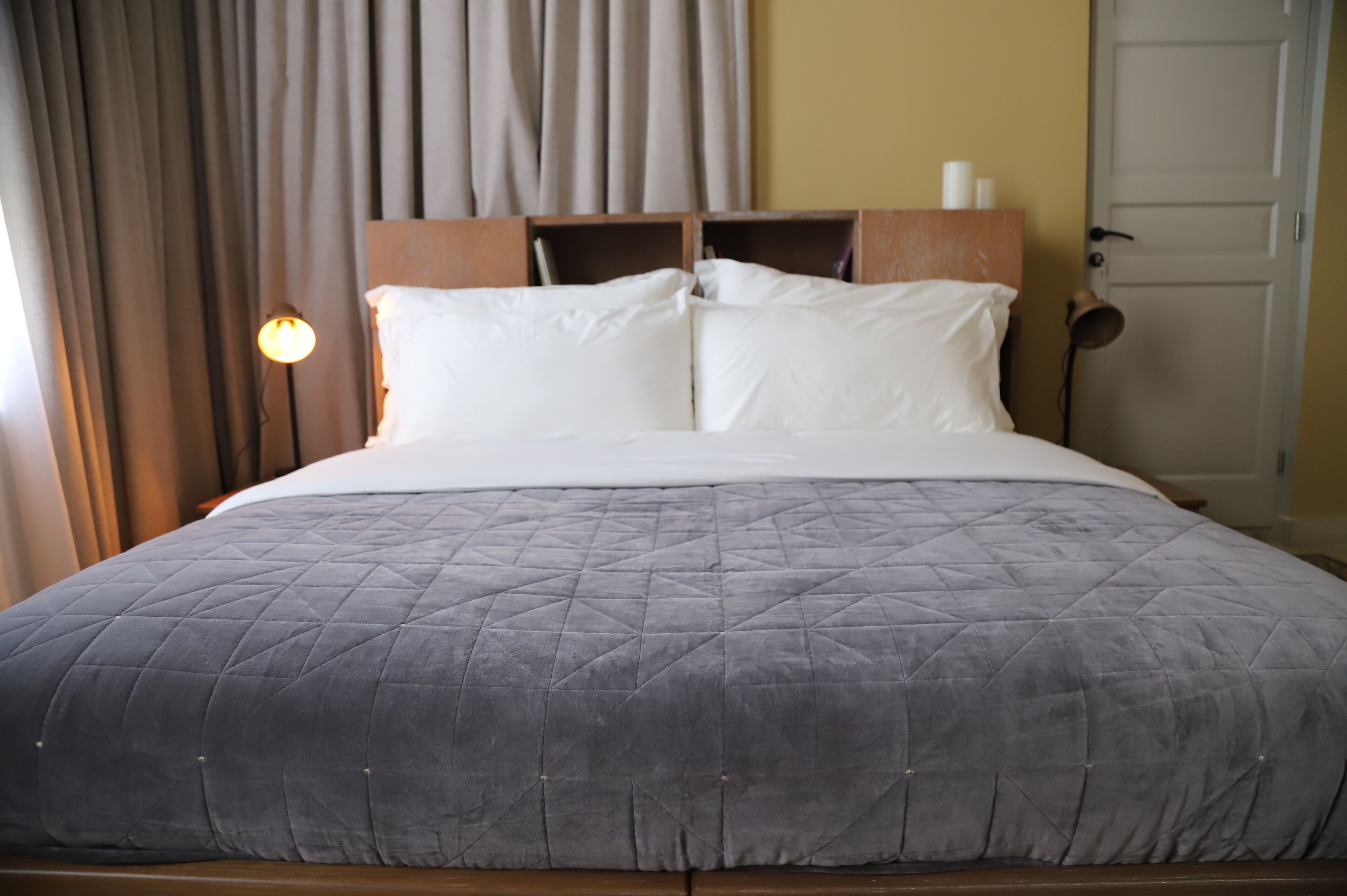A neatly made bed with a grey quilted bedspread and white pillows, a wooden headboard, and a bedside lamp.