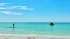 Une plage avec une eau turquoise et limpide, une personne pratiquant le paddle, et un bateau au loin.
