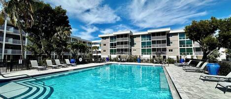 A swimming pool with lounge chairs and umbrellas, surrounded by a multi-story building with balconies.