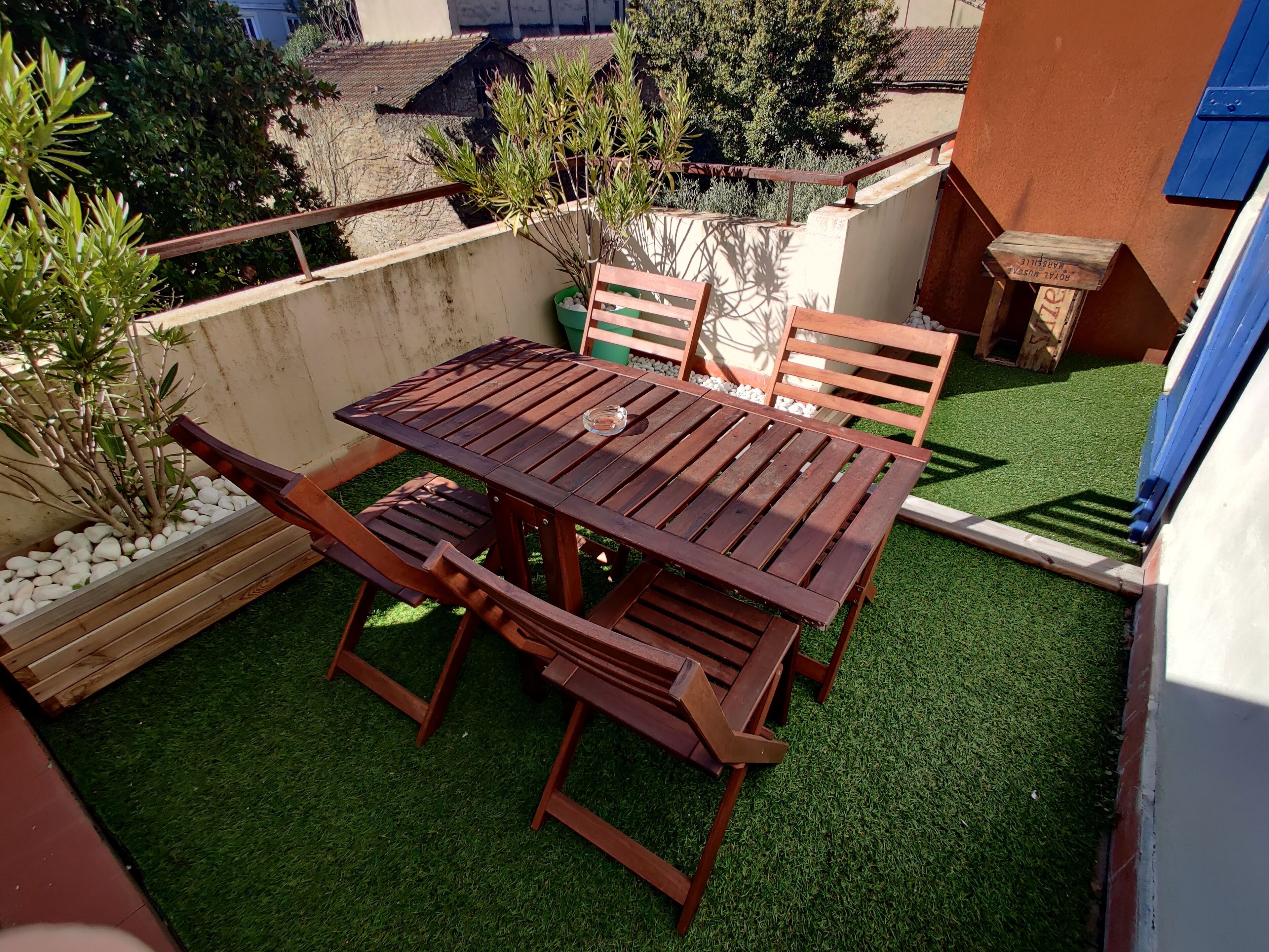 Une table et des chaises en bois sont disposées sur une pelouse artificielle, entourées de galets blancs et de plantes. Un mur en béton et une porte bleue se trouvent à l’arrière-plan.