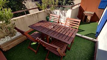 Une table et des chaises en bois sont disposées sur une pelouse artificielle, entourées de galets blancs et de plantes. Un mur en béton et une porte bleue se trouvent à l’arrière-plan.