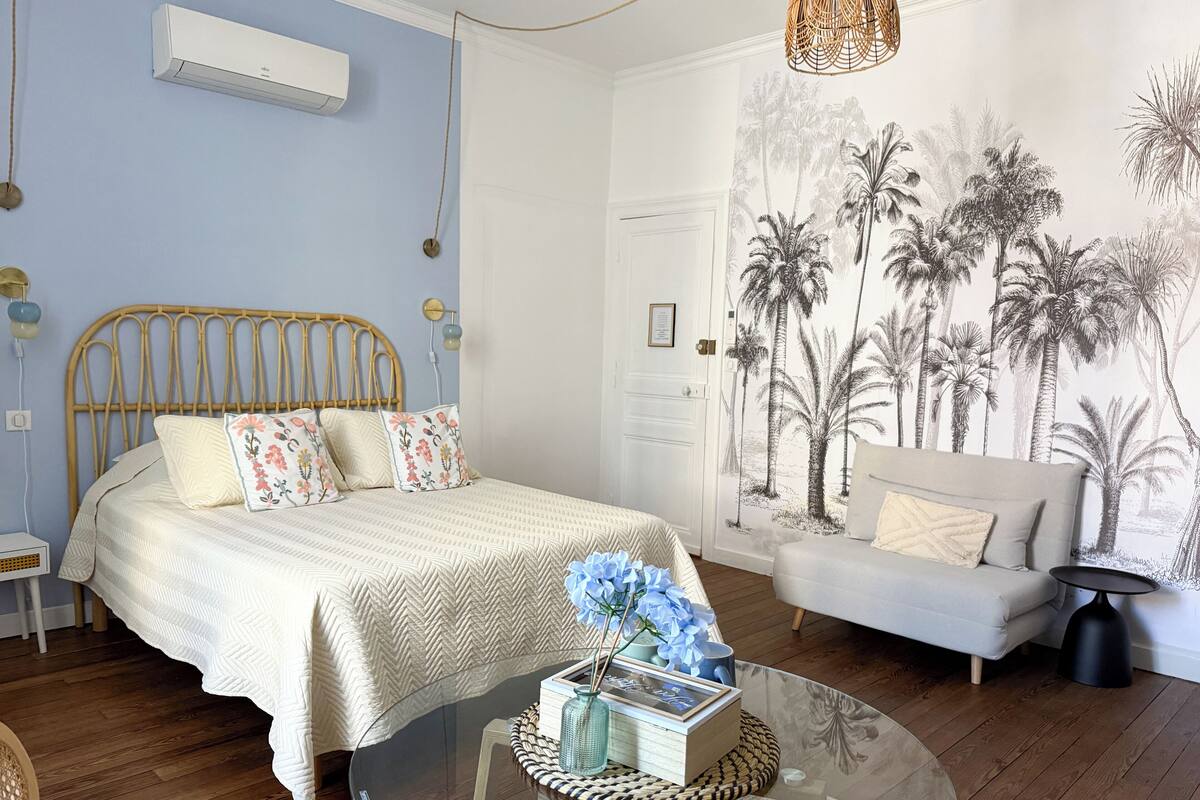 A bedroom with a bed, a sofa, a glass coffee table, and a wall with a tropical mural.