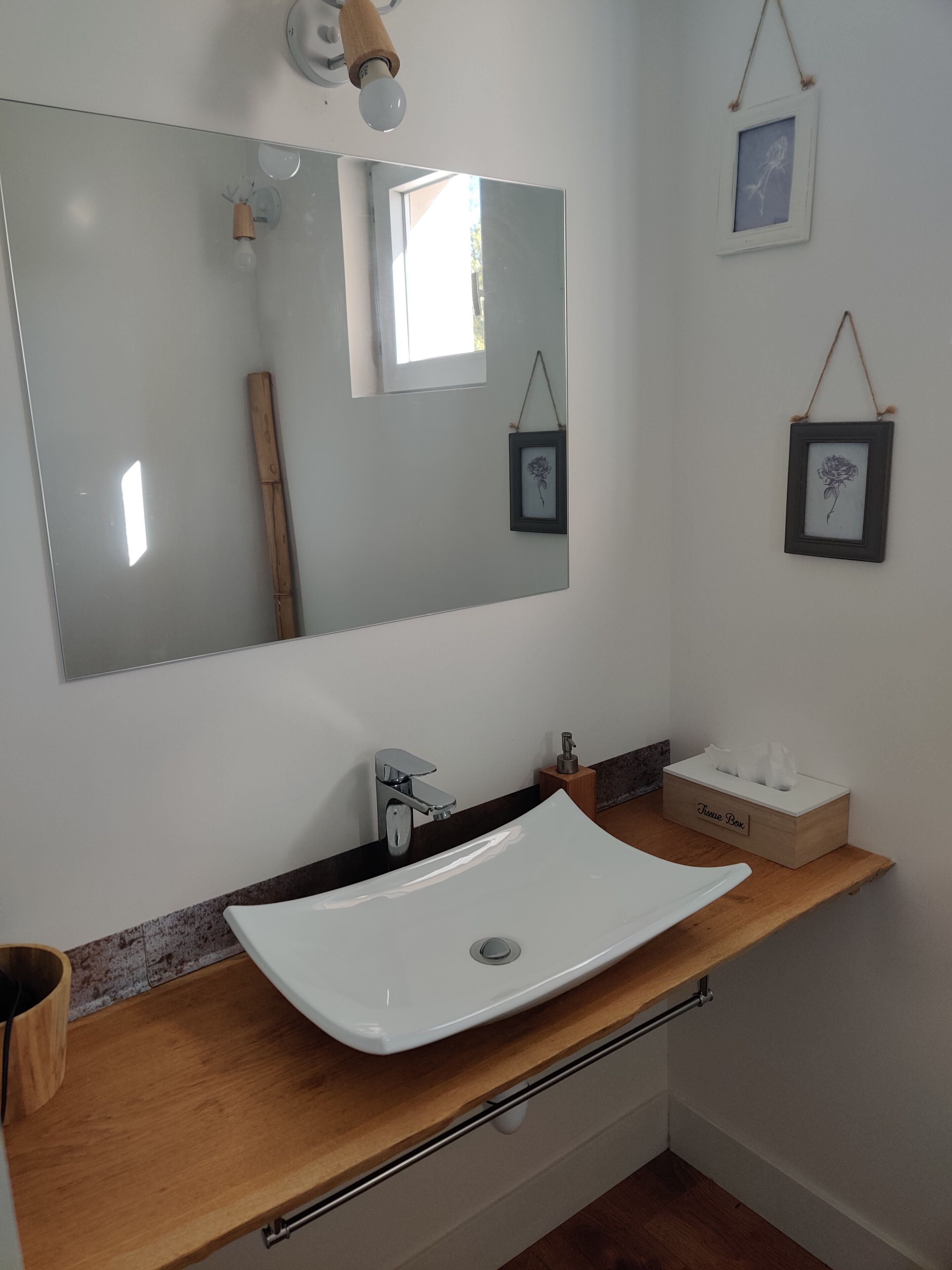 A bathroom with a white sink, wooden countertop, and a large mirror. There are two framed pictures on the wall, a tissue box, and a wooden towel rack.