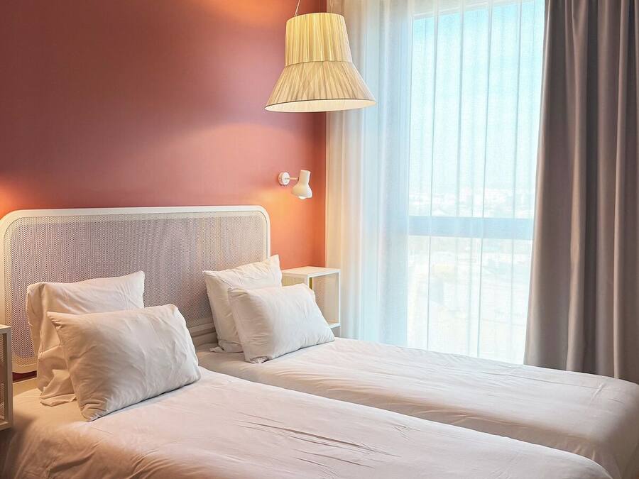 A hotel room with two beds, a large window with curtains, and a hanging lamp.