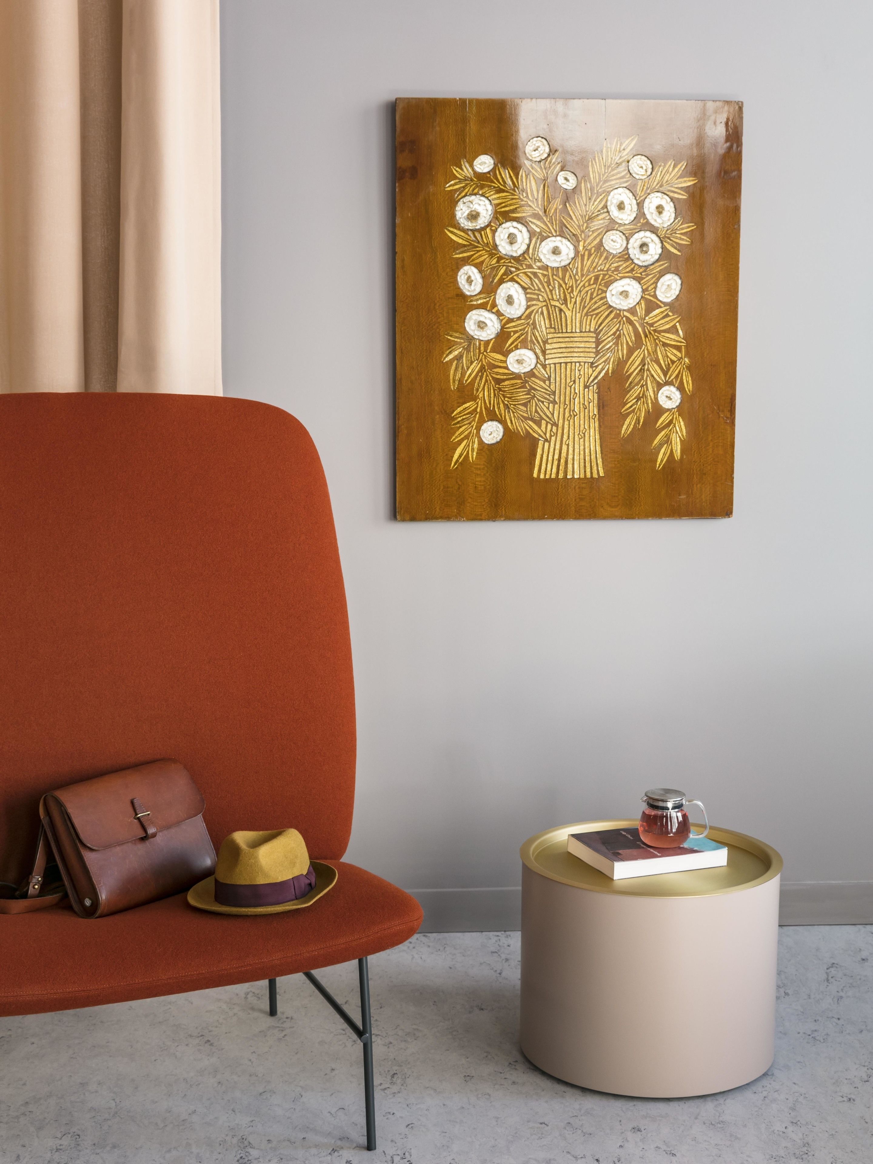 Un salon moderne avec un fauteuil rouge, une petite table ronde sur laquelle se trouvent un livre et une théière, et une œuvre d’art murale représentant un arbre en fleurs.