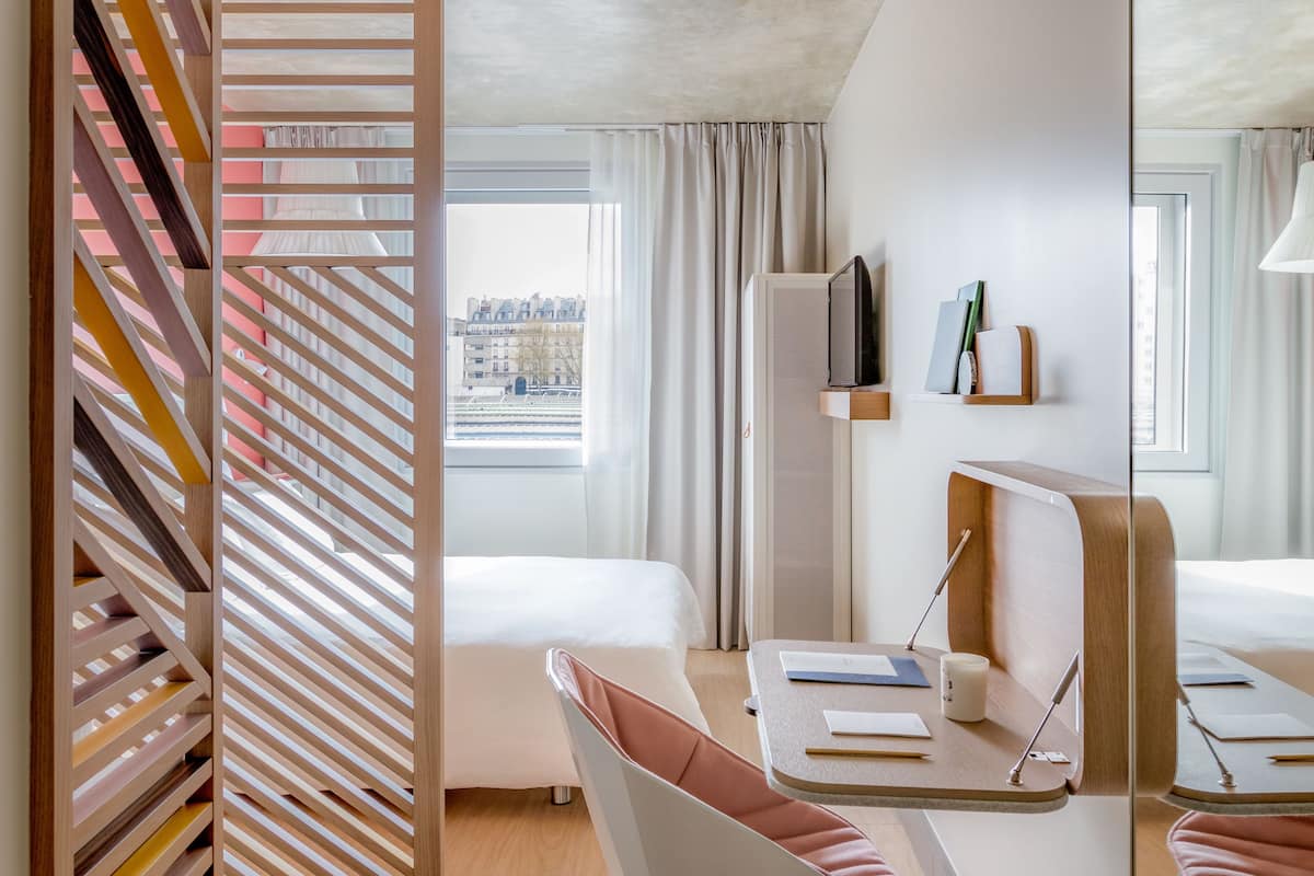 Una habitación de hotel moderna con un ventanal grande, un escritorio con silla, una cama y un panel decorativo de madera.
