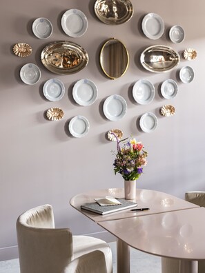 Un comedor con una mesa redonda, sillas blancas, un florero con flores y una pared decorada con platos y un espejo.
