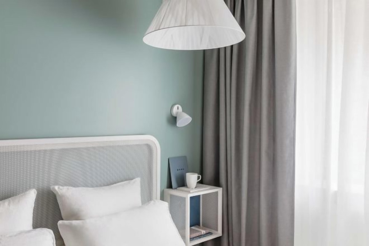 A neatly arranged hotel room with a bed, a bedside table with a lamp, and a small shelf with a book and a cup.