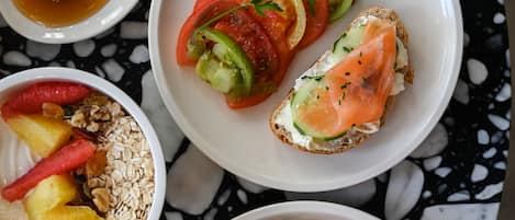 Un desayuno con una variedad de alimentos, incluyendo un plato con rodajas de tomate y pepino, y una rebanada de salmón ahumado sobre tostada, un bol de yogur con frutas y granola, y un plato con cruasanes.
