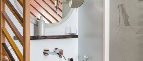 Un baño con un lavamanos blanco, un espejo redondo sobre un fondo a rayas, una puerta de listones de madera y una pared de mármol.