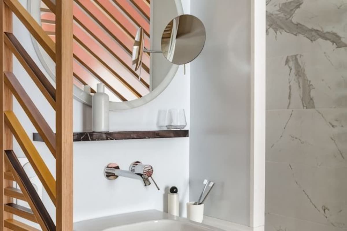 Une salle de bain avec un escalier en bois, un miroir rond, un lavabo blanc et un mur en marbre.