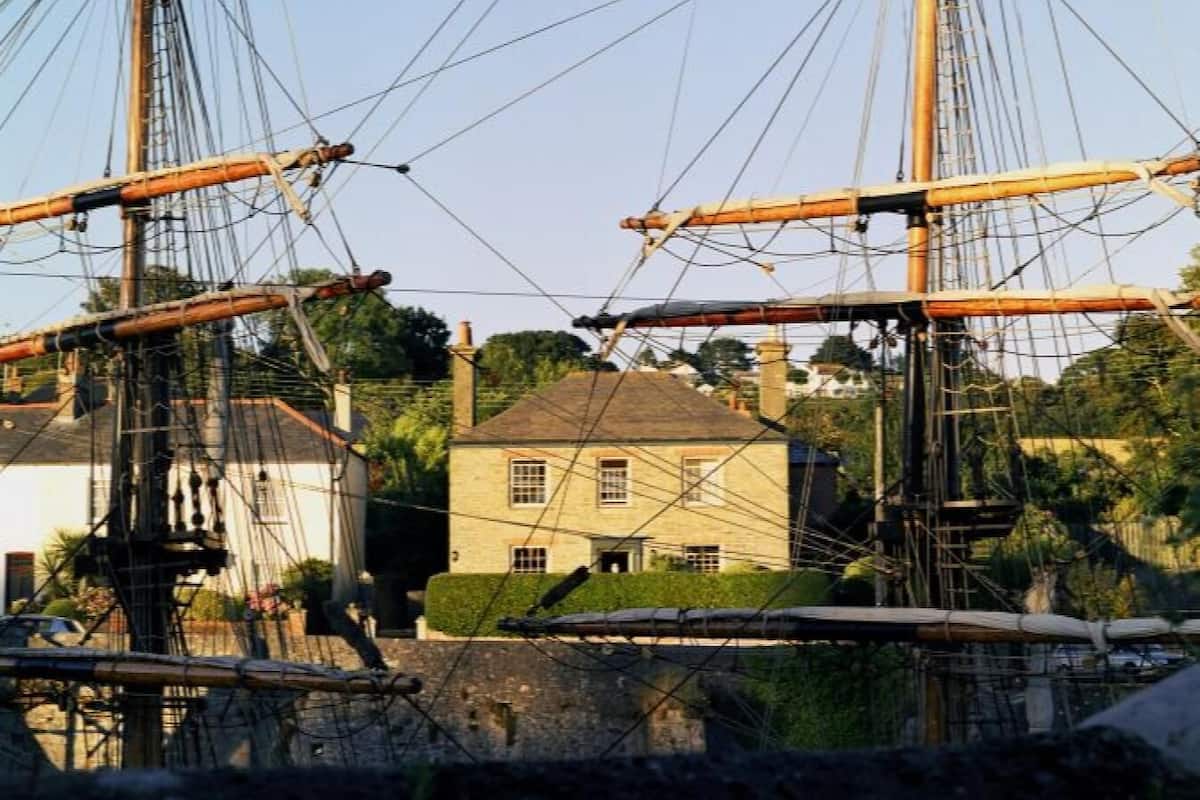 A historic ship with masts and rigging, docked near a house with a gabled roof and multiple windows.