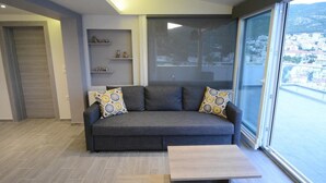 A modern living room with a grey sofa, patterned cushions, a wooden coffee table, and a large window offering a city view. - "VERDE BLUE" – Stylish Loft with Panoramic Terrace in Kavala (Kavala)