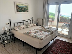Ein ordentlich bezogenes Bett mit einem gemusterten Überwurf, ein kleiner Nachttisch mit Lampe, ein Bild an der Wand und ein Blick ins Freie durch eine Schiebetür.