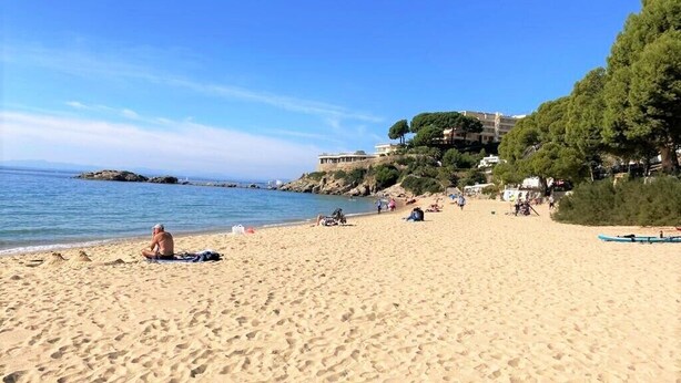 A sandy beach with footprints, a few people, and a building with trees on a hill in the background.