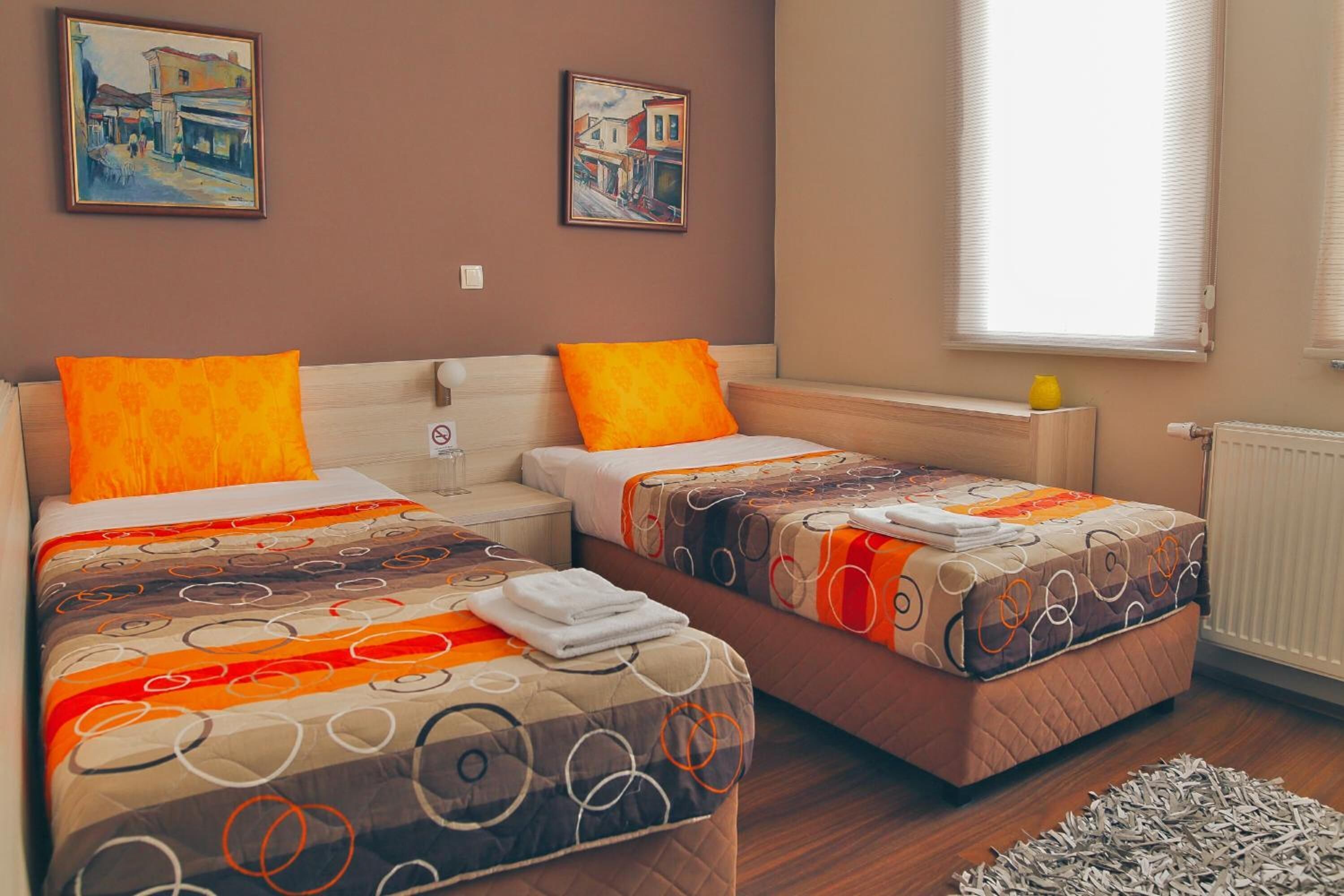 A hotel room with two beds, each with an orange pillow, a patterned bedspread, and a small table with a yellow vase.
