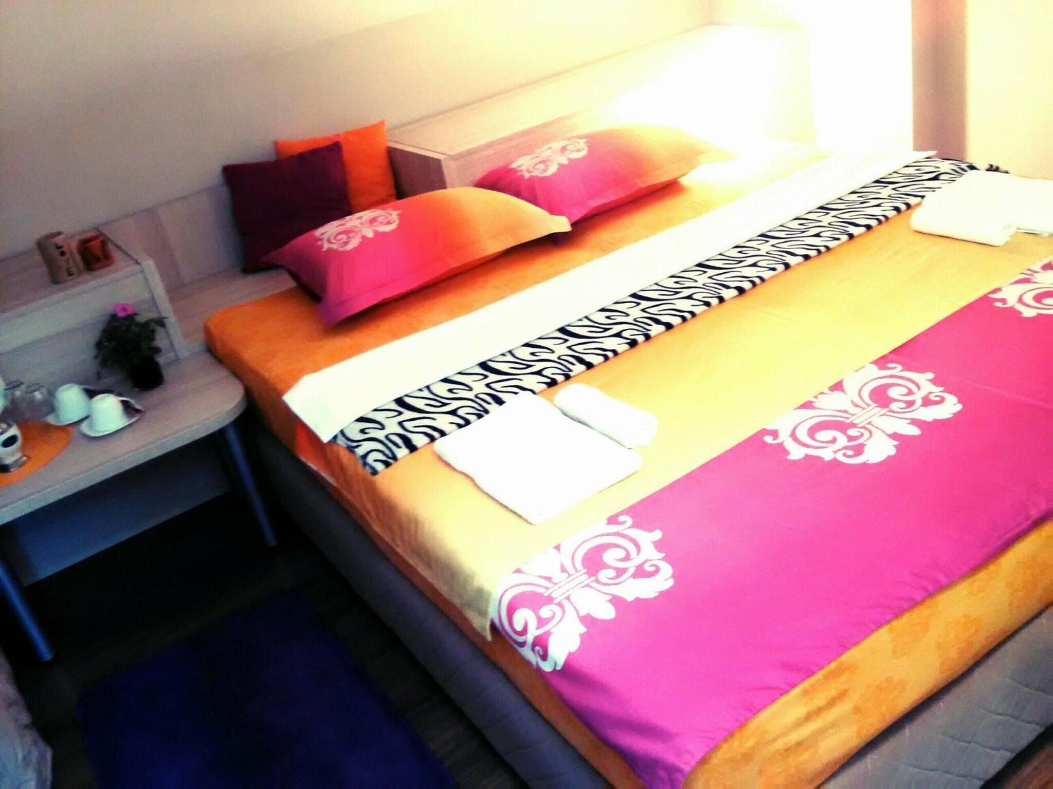 Cama bem arrumada com colcha estampada, travesseiros e mesa de cabeceira com abajur e uma pequena planta.