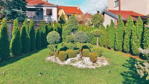 A well-maintained garden with neatly trimmed hedges and a central tree, surrounded by a brick wall and houses with red-tiled roofs. - Hotel Bulevar (Bitola)