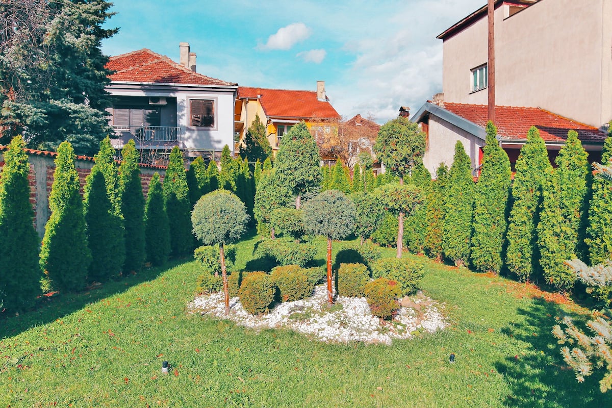 A well-maintained garden with neatly trimmed hedges and a central tree, surrounded by a brick wall and houses with red-tiled roofs.
