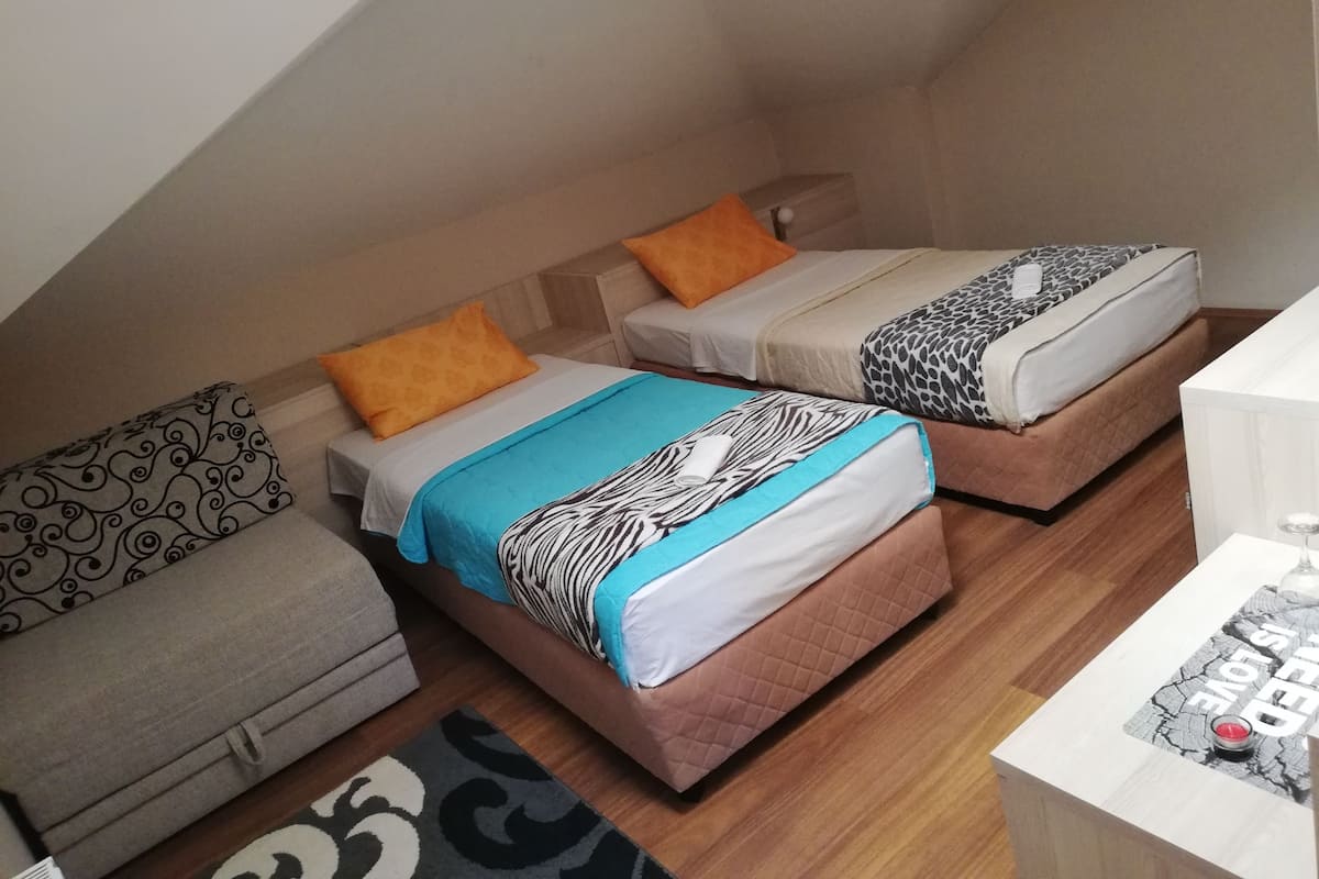 A small, cozy room with three beds, each with different patterned bedding. There is a sofa and a small table with a decorative item on it.