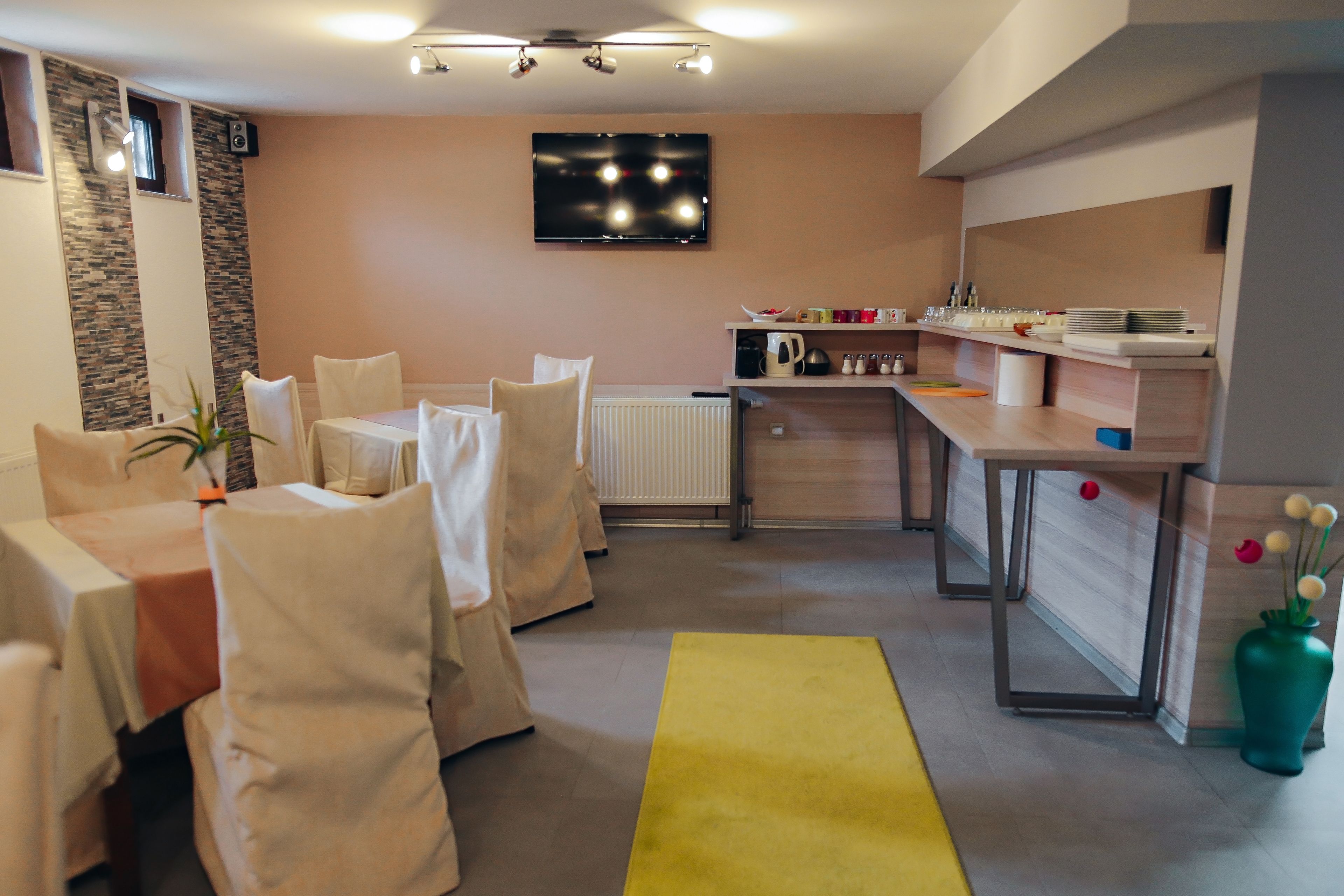 A modern dining area with a kitchen in the background, featuring a flat-screen TV, a dining table set with chairs, and a yellow rug.